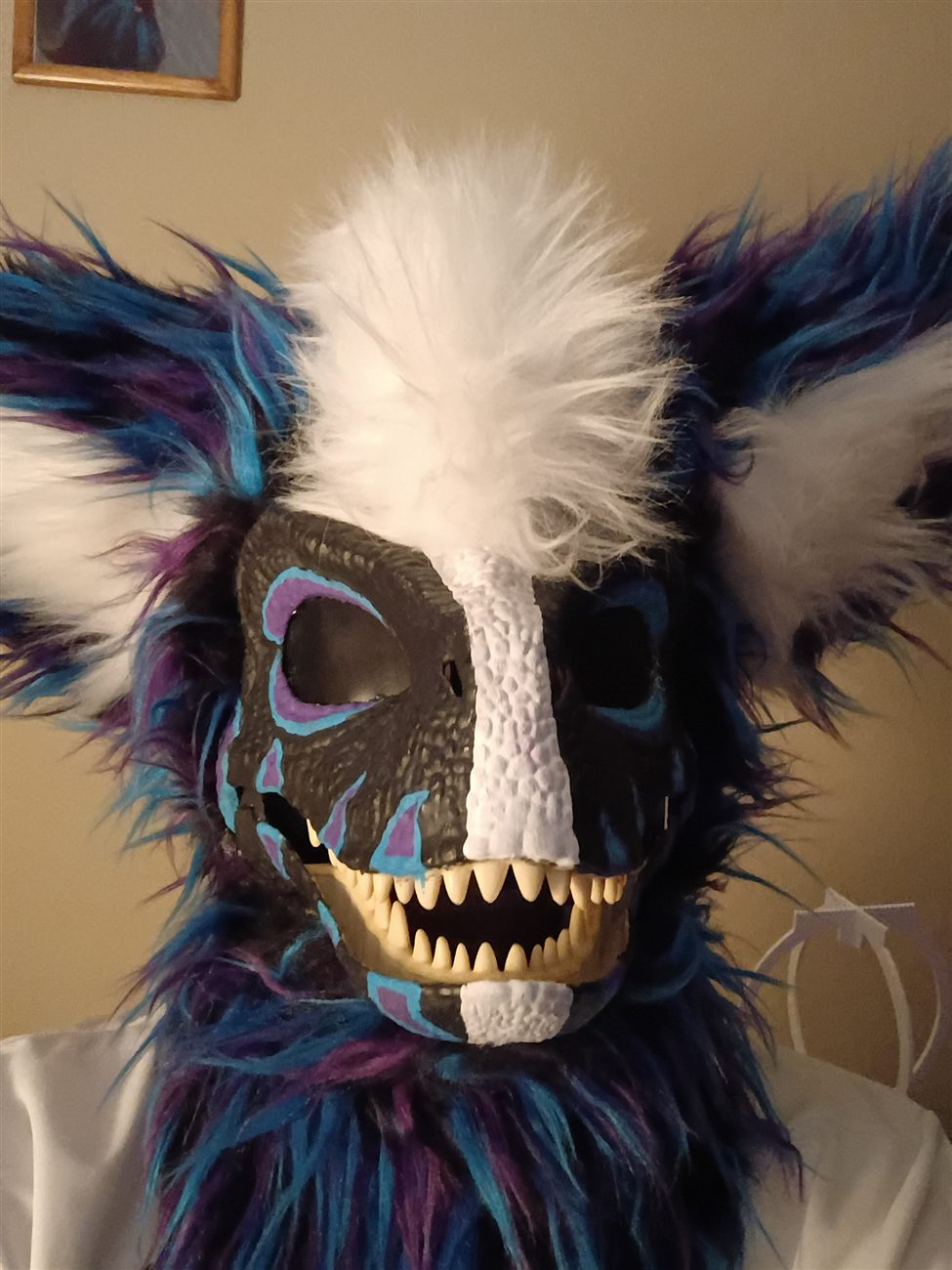 A blue, purple, and black custom fursuit head in progress, with mesh eyes and painted fangs