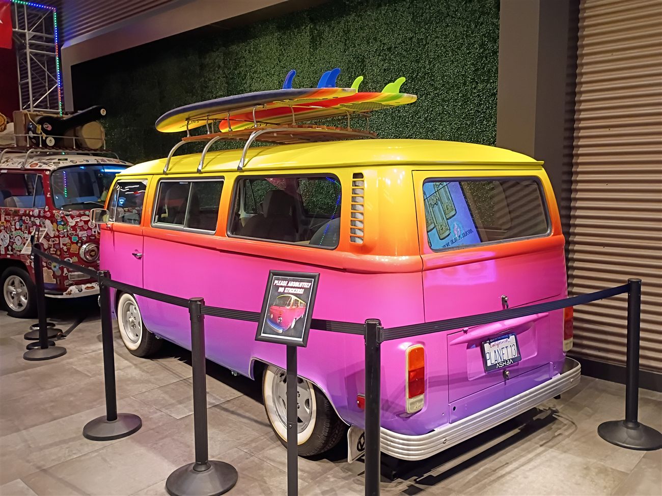A sunset-gradient VW bus with surfboards on the roof rack and a Planet 13 license plate